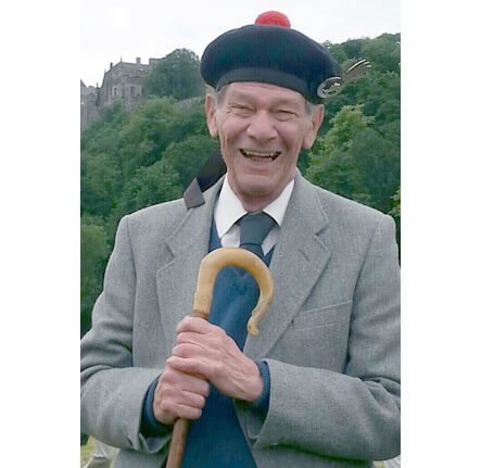 A photograph of Alexander Gilbert Haldane Robertson - The chief of Clan Donnachaidh. He wears his Scottish tartan kilt with white collared shirt and grey tweed kilt jacket, stood smiling with foliage of green trees behind him in the background.